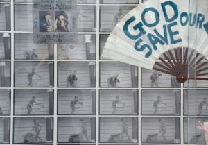 Fan with God Save our NHS painted on it hung in front of wall paper of images of nude woman doing tasks