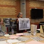 Woman holding up a newsprint poster behind table covered in slide files, magazines and newsprint publications surrounded by workshop participants.