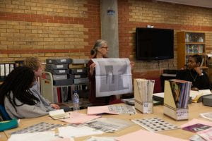 Woman holding up a newsprint poster behind table covered in slide files, magazines and newsprint publications surrounded by workshop participants.