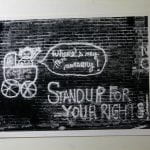 Black and white photograph of hand painted graffiti on a wall, shows a baby in a pram with speech bubble, text reads: where is my free nursery? Stand up for your rights!