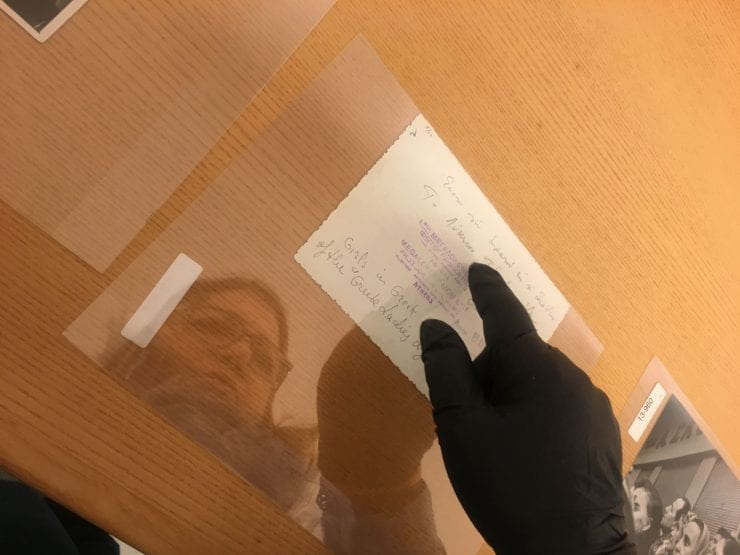 The surface of a table, in the reflection a woman's face stares towards a white sheet with handwritten text. Her hand in a black glove points to the script.