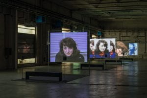 Installation view of four screens in a large concrete gallery room, each screen displaying images of a woman 