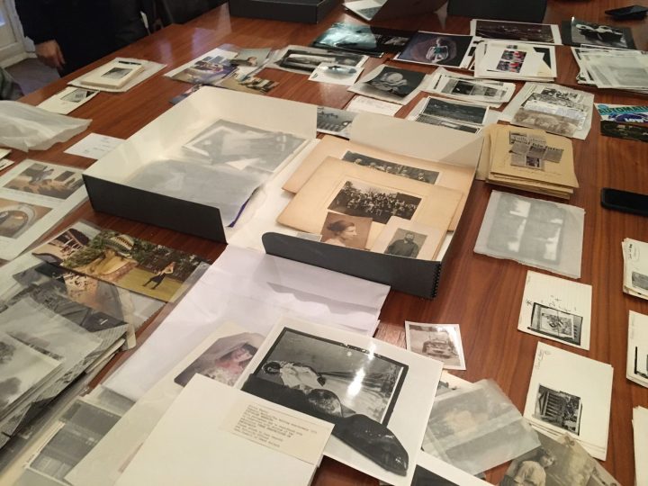 Across a large table an archive box has been opened and the photographs, postcards and other printed matter it contained have been laid out and roughly organised.