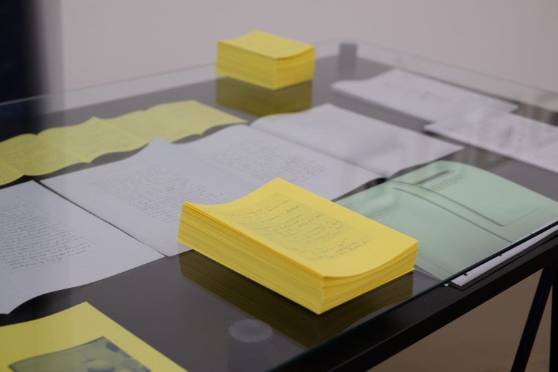 On a glass topped vitrine white, light yellow and light green papers are laid out. Stacks of bright yellow paper booklets sit on top of the glass. 