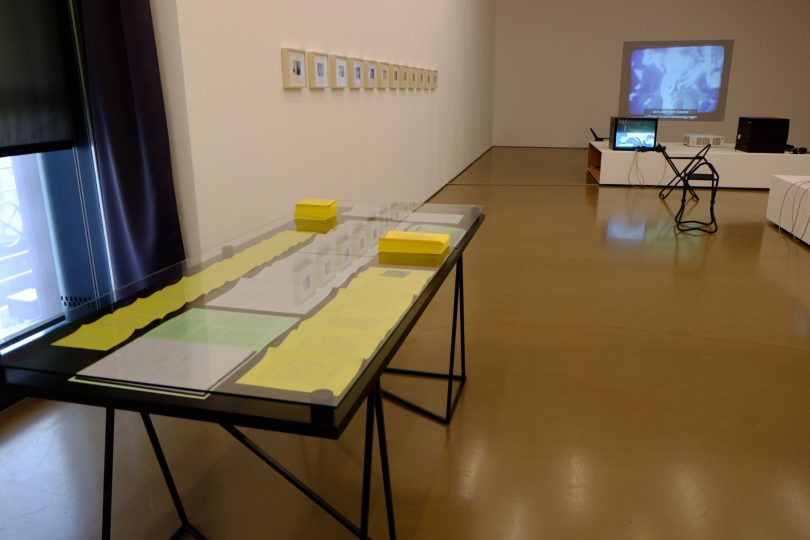 In the foreground of a gallery space is a glass vitrine table with papers spread out inside and stacked on top. Nearby on the wall is a series of small, framed photos and further back in the room is a square television monitor.