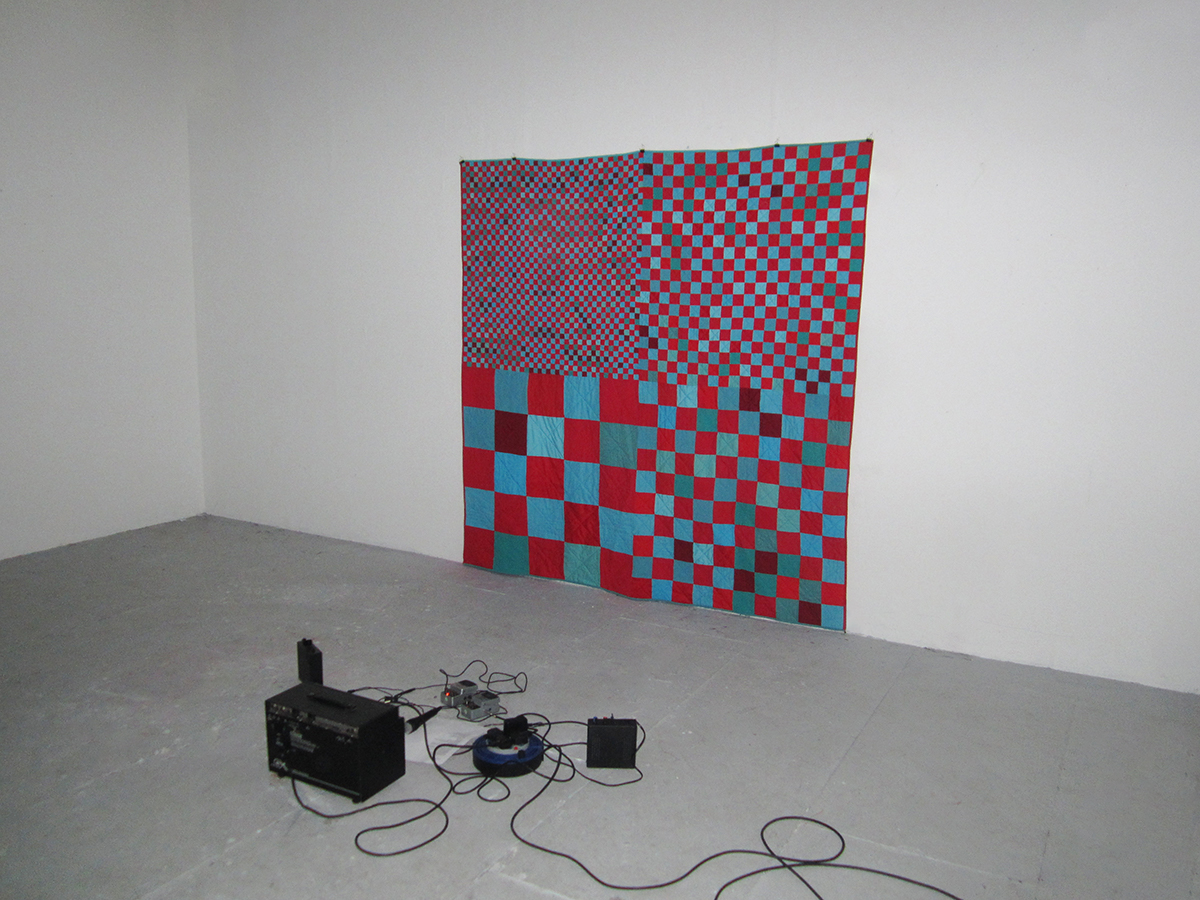 Photo of A red and blue checkered quilt hanging on a white wall, with audio equipment on a grey floor.