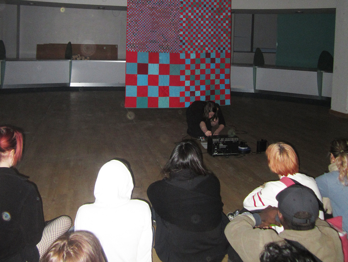 A photo from the audience of a performance in front of a red and blue checkered quilt. 