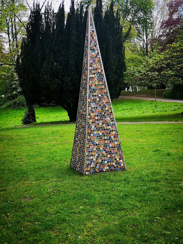 A photo of an elongated pyramid made of used coffee capsules, standing in a field.