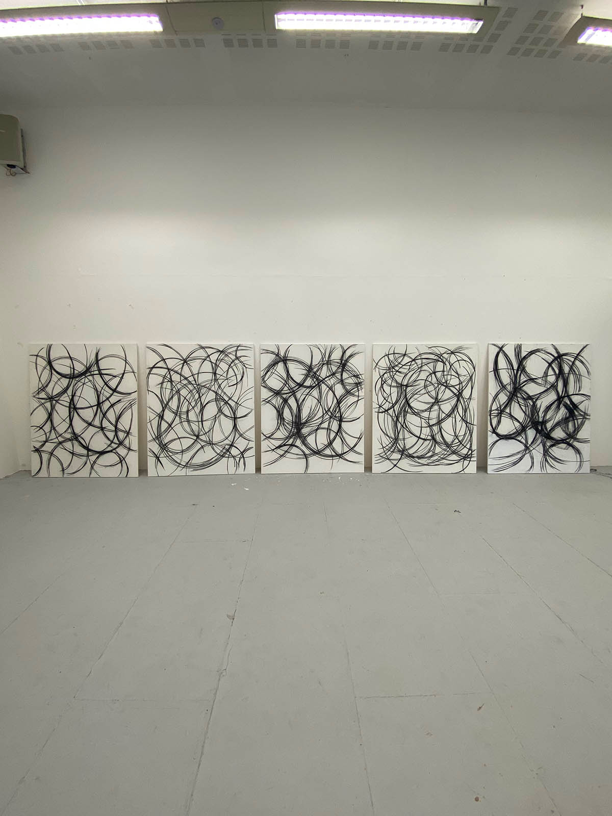 A picture of five canvases standing against a wall, each covered in charcoal-drawn swirls.