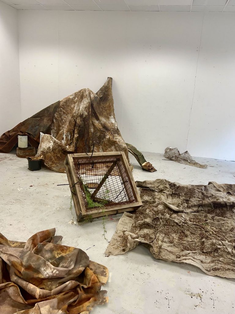A photo of an installation in a room consisting of brown-stained material draped over hidden objects.