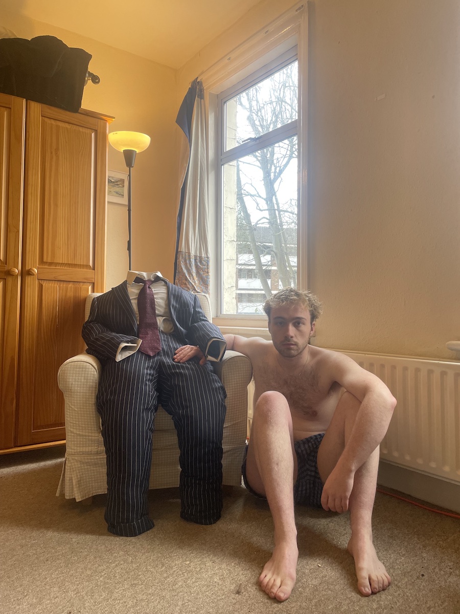 A photo go a man sitting in pants on the floor, and a suit sitting empty and upright in an armchair next to him.