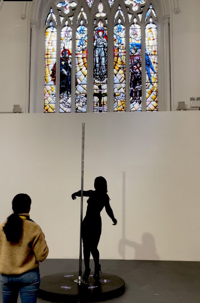 A figure in a black unitard pole dancing beneath a stained-glass window.