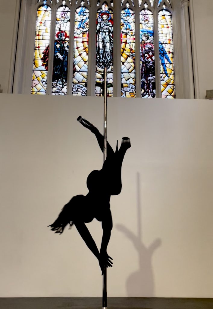 A figure in a black unitard pole dancing beneath a stained-glass window.