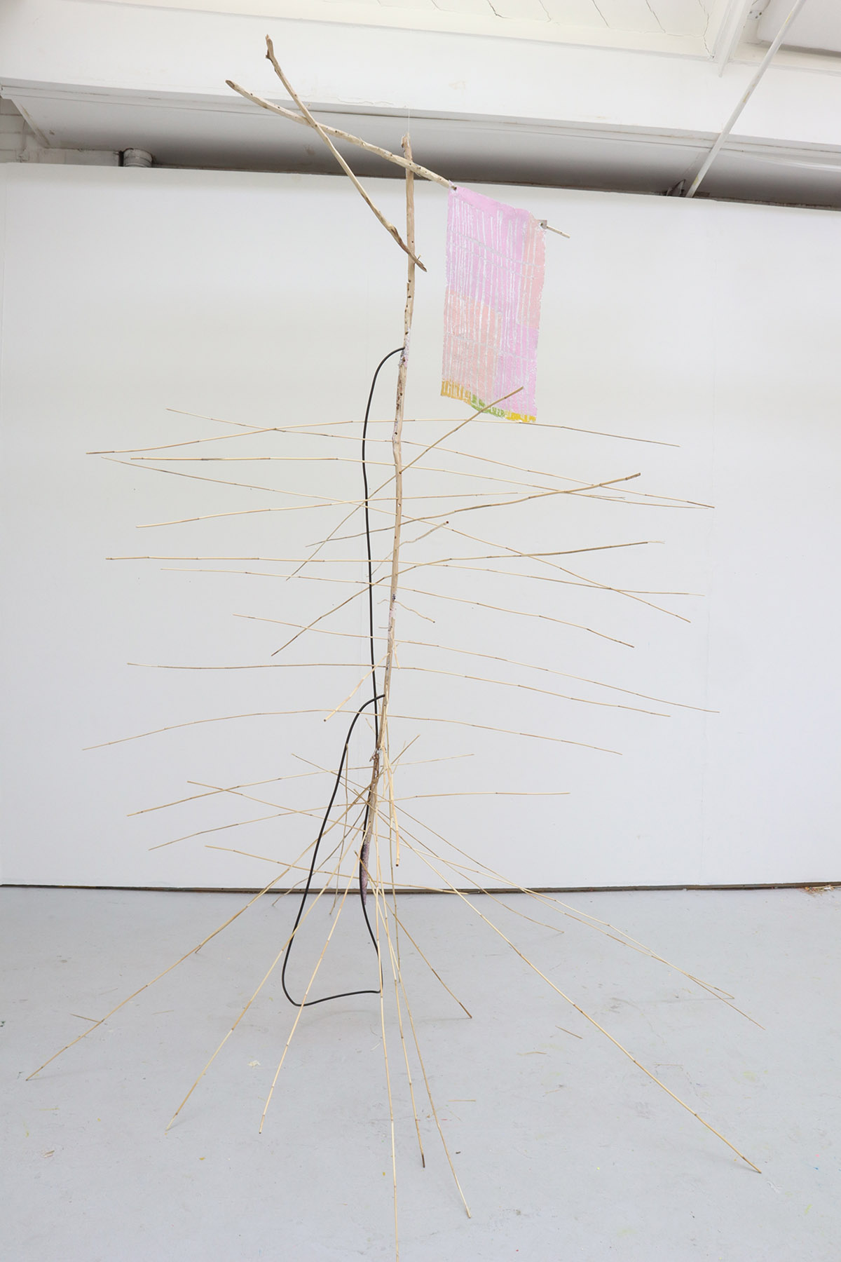 A freestanding sculpture made of thin sticks, from which hangs a pink oil pastel drawing with silver tape running through it.