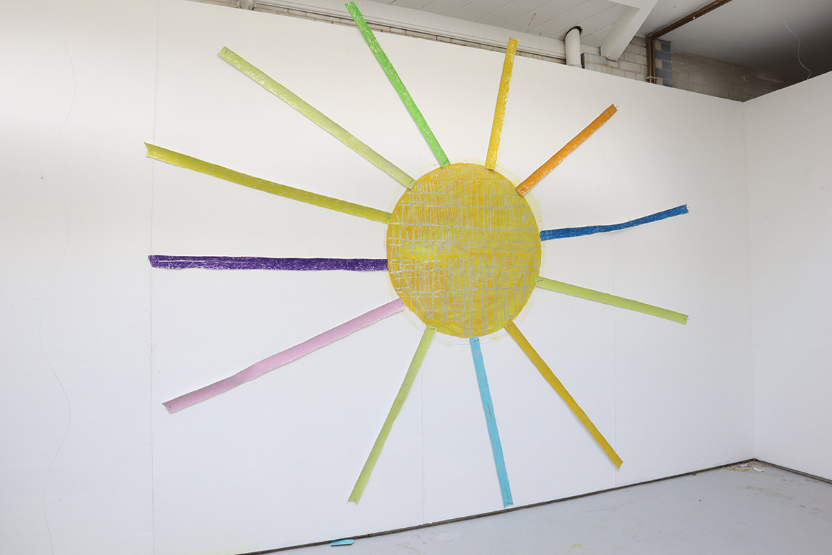 A colourful oil pastel drawing of a sun shape with a silver tape running through it, hanging on a white wall.