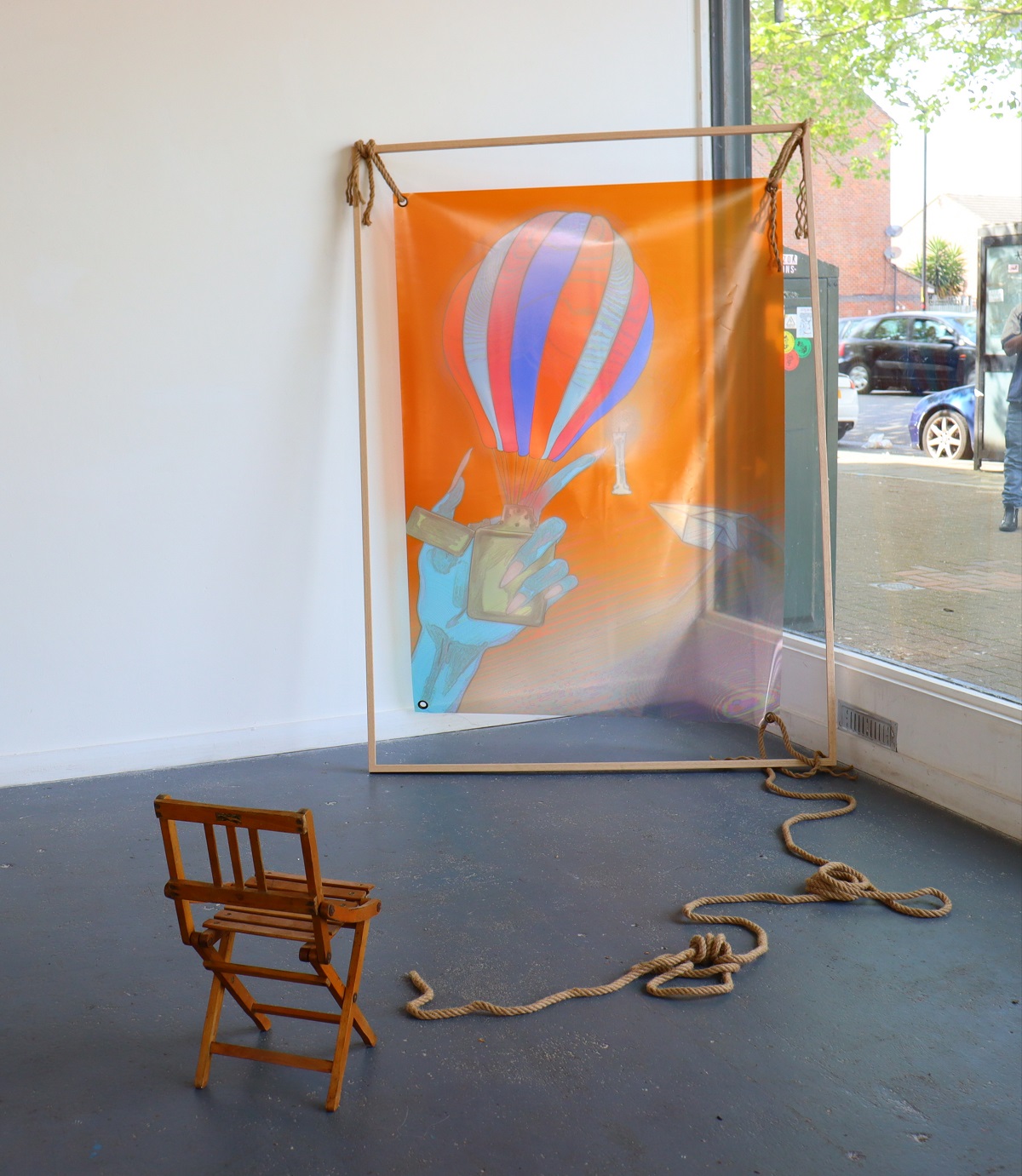 A picture of an installation where a small wooden chair is facing a colourful acetate print hanging from an oak frame via ropes.