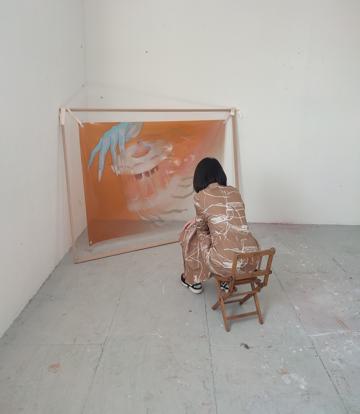 A picture of an installation where someone sitting in a small wooden chair is facing a colourful acetate print hanging from an oak frame via ribbons, in the corner of a room.