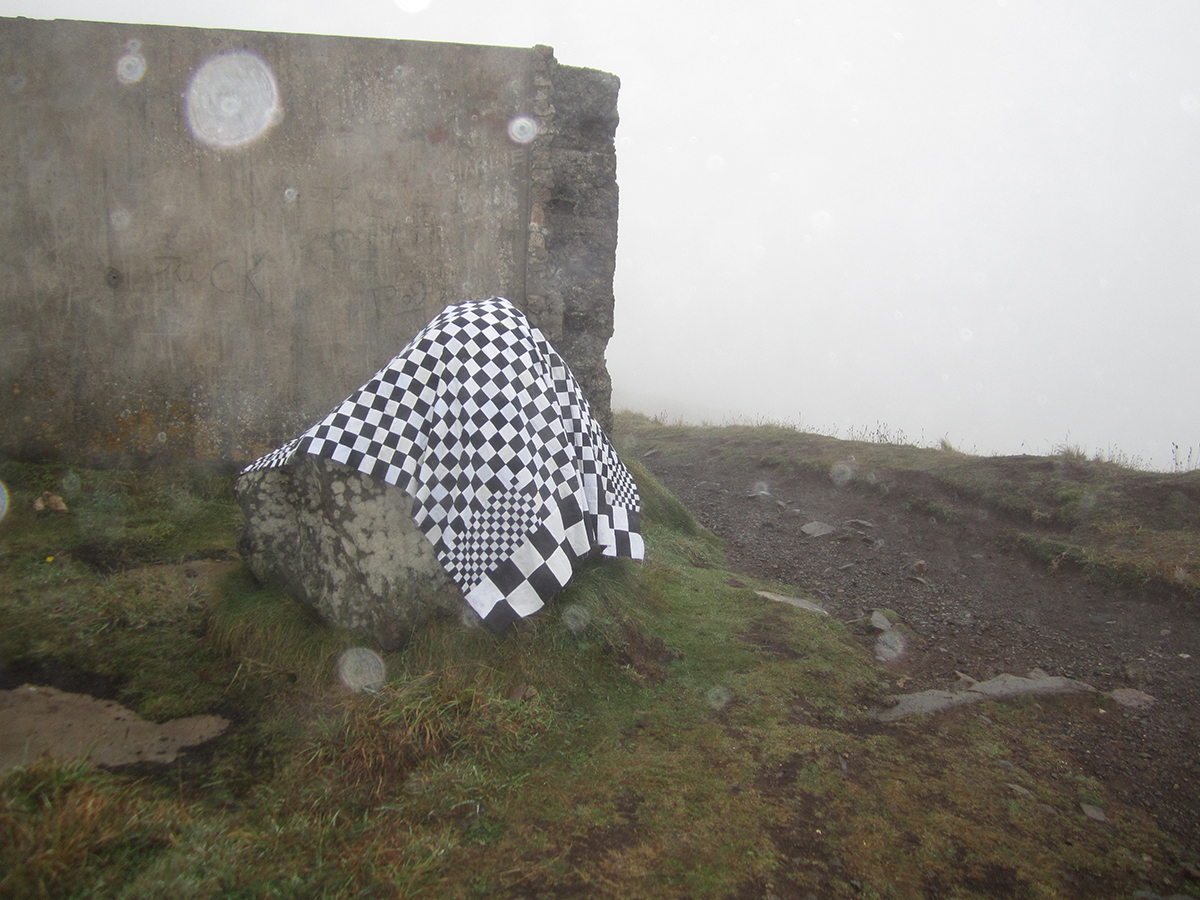 A photo of a black and white checkered quilt hanging over a rock on a rainy moor.