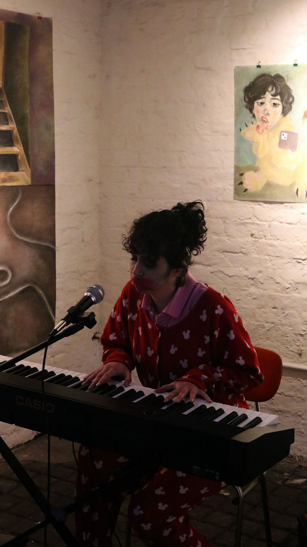 Photo of a girl sitting in a red onesie, playing a piano in front of some paintings.