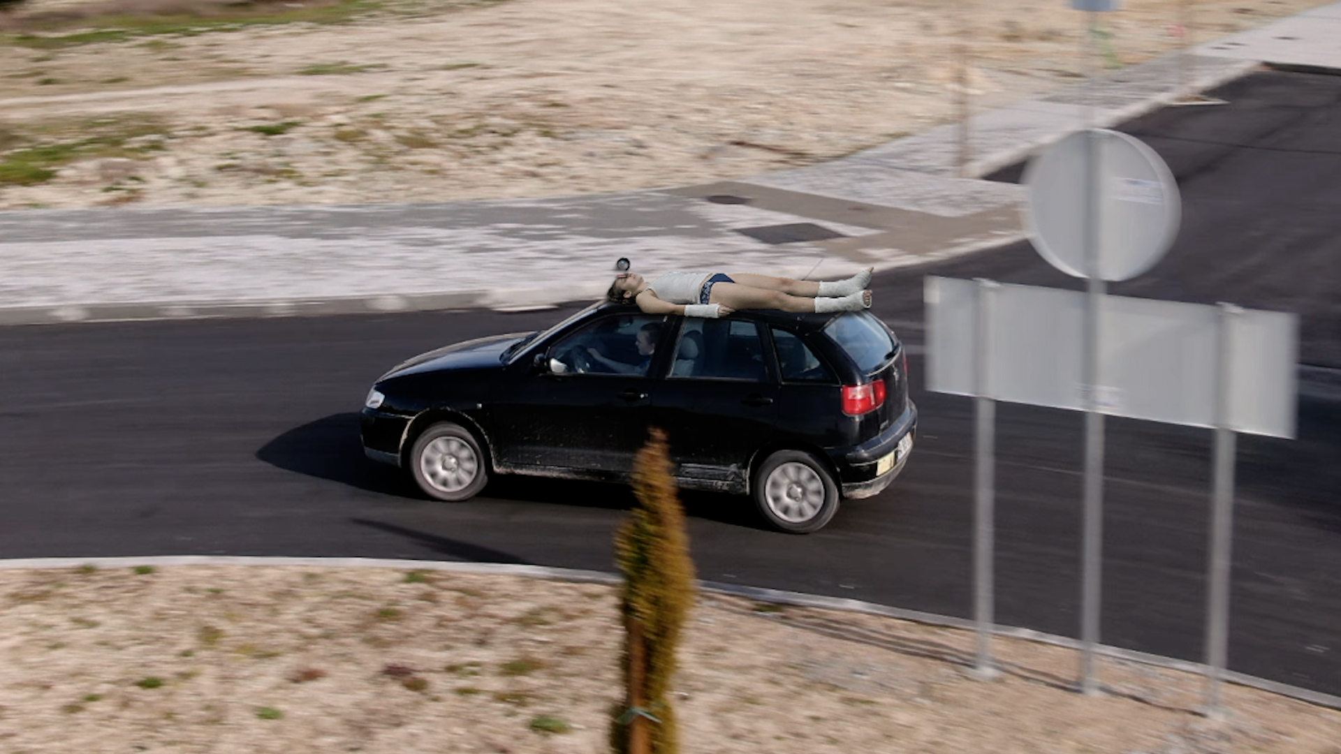 A shot of a car driving with a person laying on their back on the top of the vehicle.