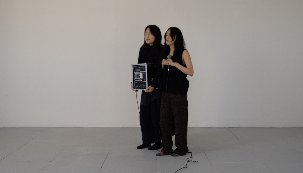 A photo of two people standing side by side in a room. One holds a microphone which is connected to an amp the other person is holding.