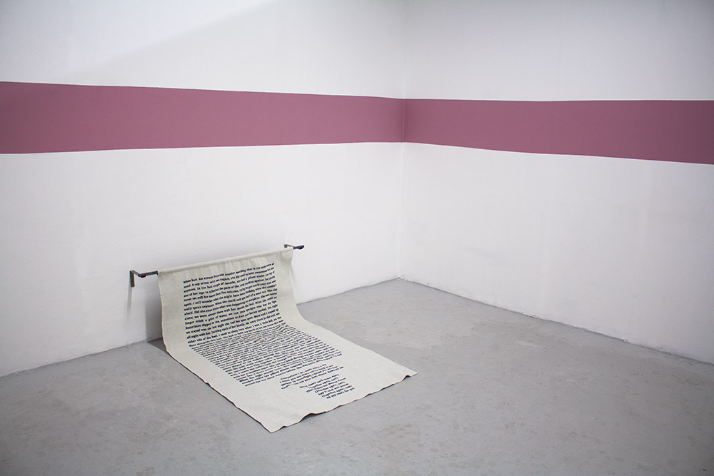 A rectangular piece of fabric, with embroidered song lyrics, hangs from a metal pole on the wall, folding onto the floor. A pink stripe of paint runs above it.