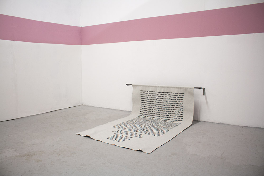 A rectangular piece of fabric, with embroidered song lyrics, hangs from a metal pole on the wall, folding onto the floor. A pink stripe of paint runs above it.
