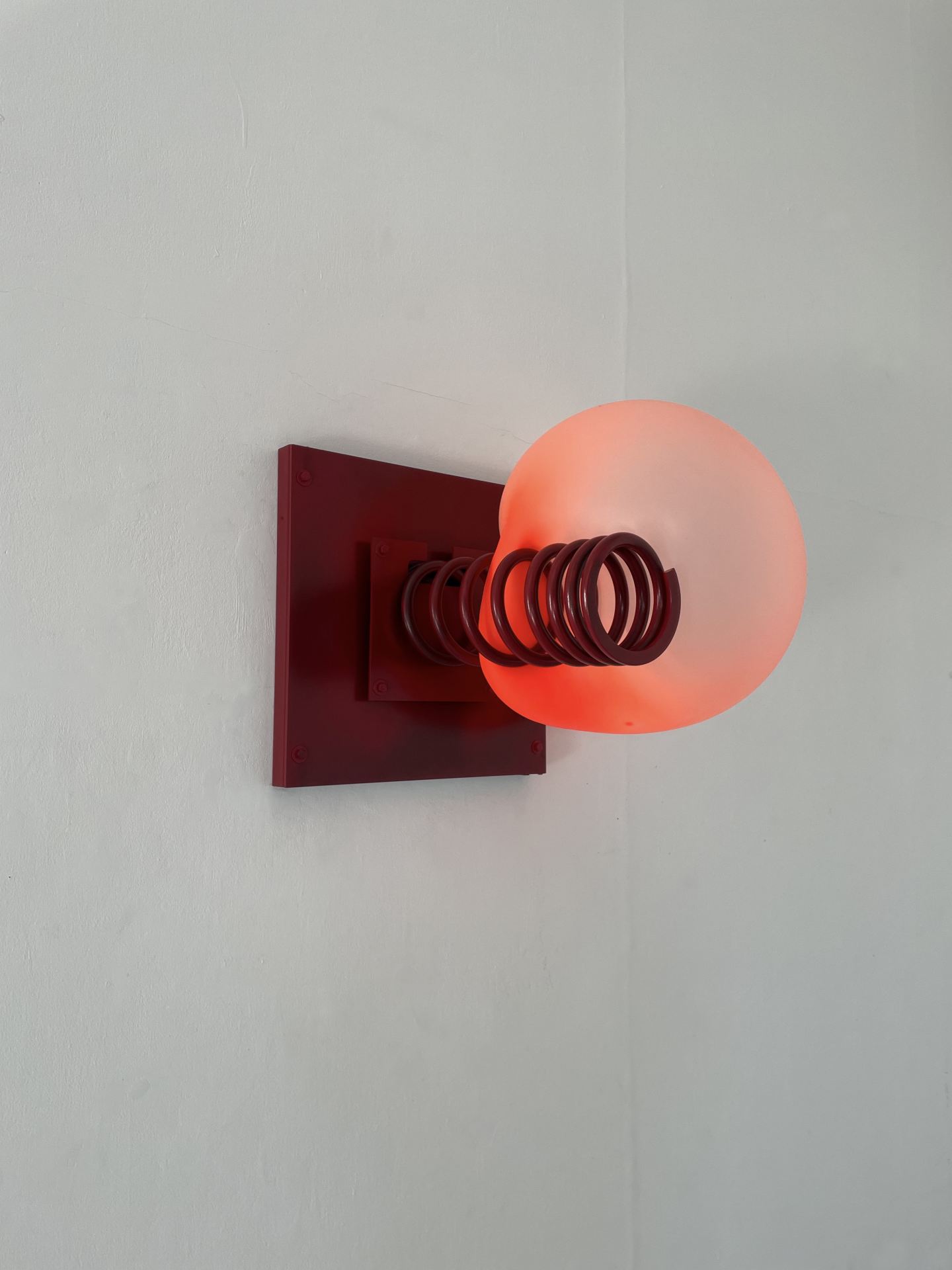 A sculpture comprised of a red spring hanging from a red square - pinching a fleshy circle - hangs from a white wall.