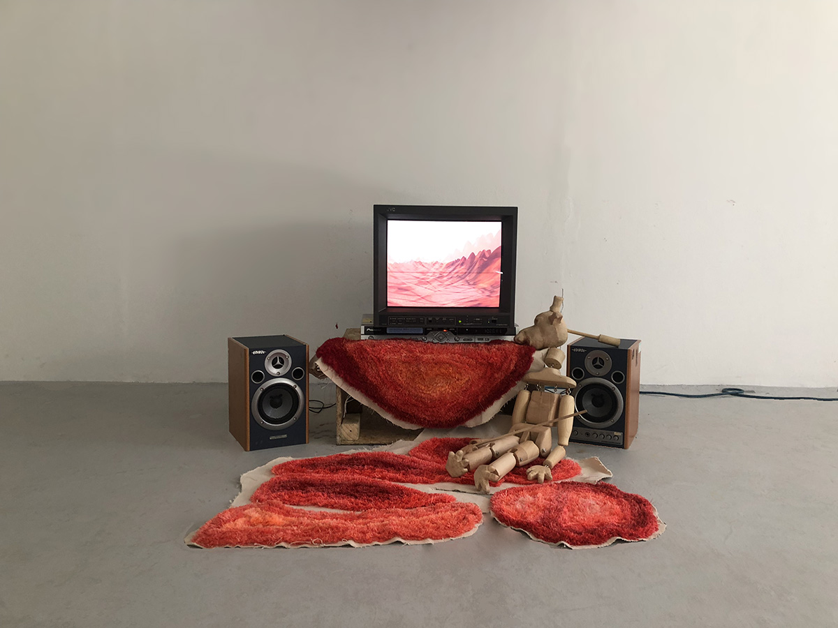 A wooden puppet is sitting on an orange tufted rug, next to two speakers and a television showing a red landscape.