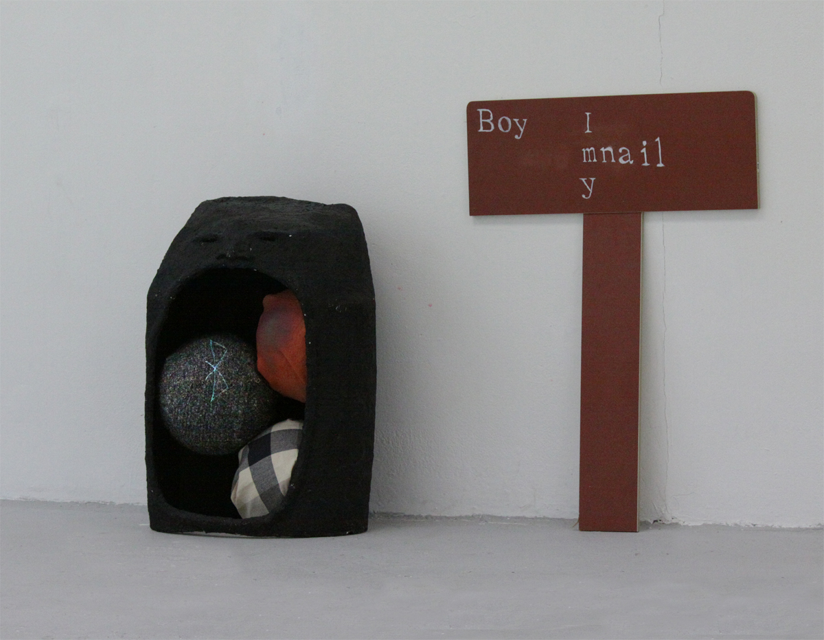 A sculpture shaped like a large head holds 3 footballs in its mouth. Next to it stands a small wooden sign reading 'Boy Imnaily'.