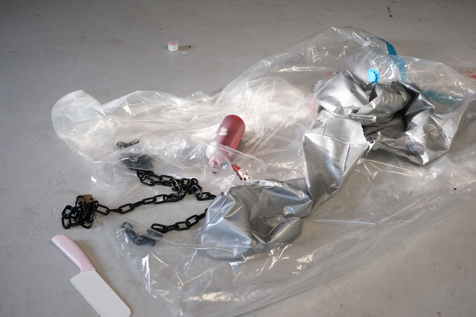Plastic wrap, a deflated mannequin, a bottle of fake blood and a black chain are in a pile on a grey floor.
