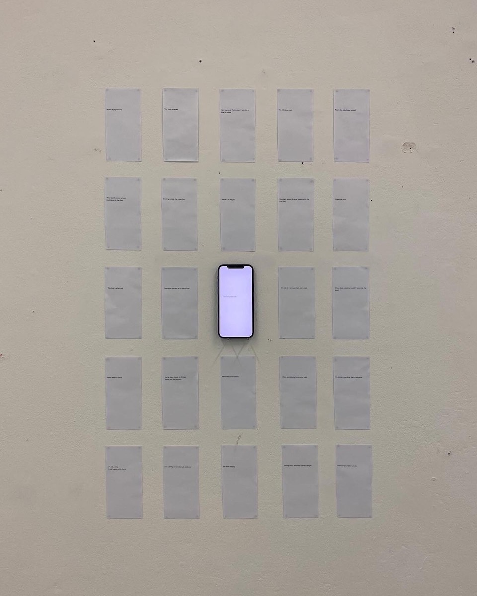An iPhone is hanging on a white wall, surrounded by 24 pieces of white paper with black writing.