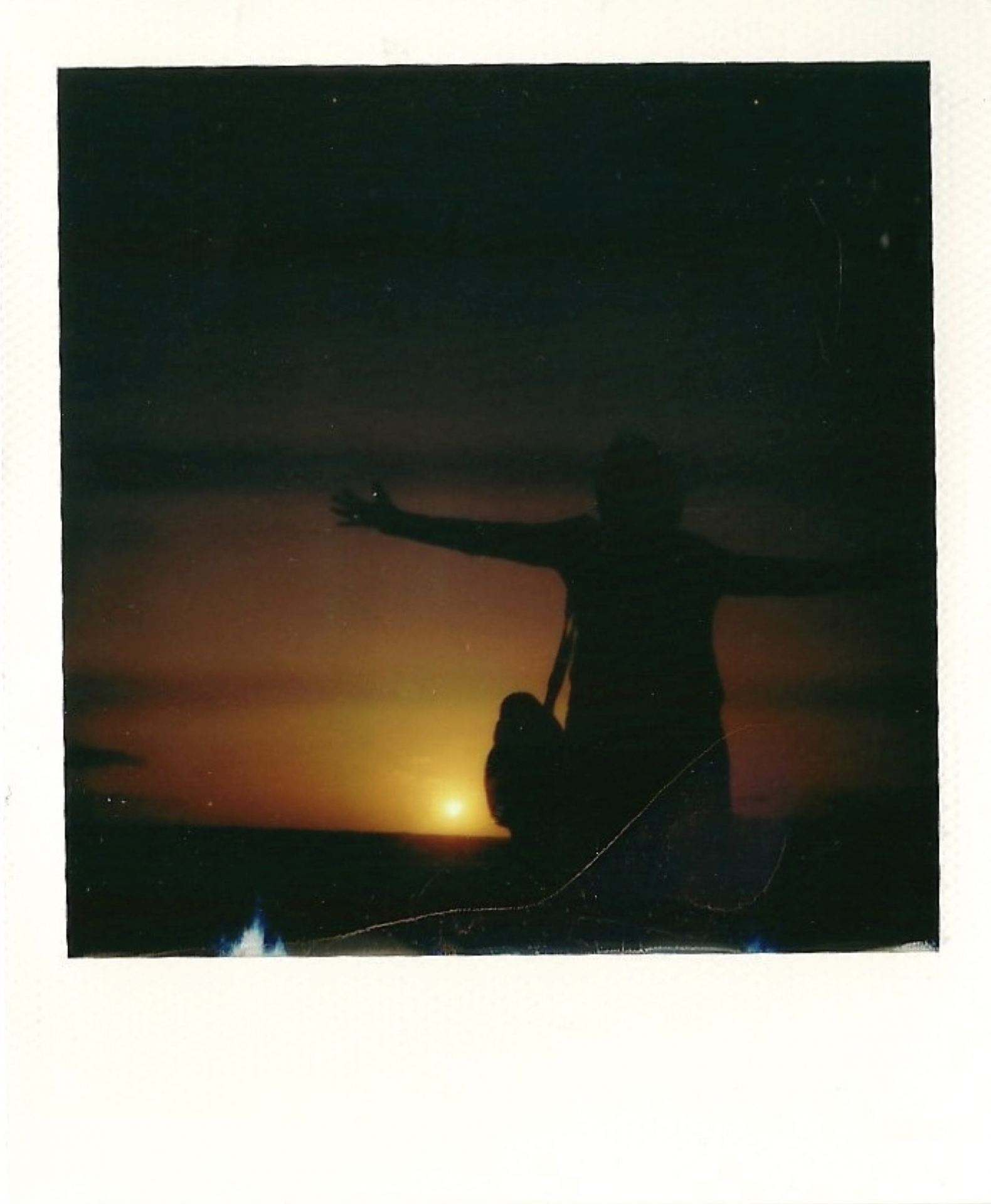A polaroid photograph of somebody standing against a sunset, with their arms outstretched.