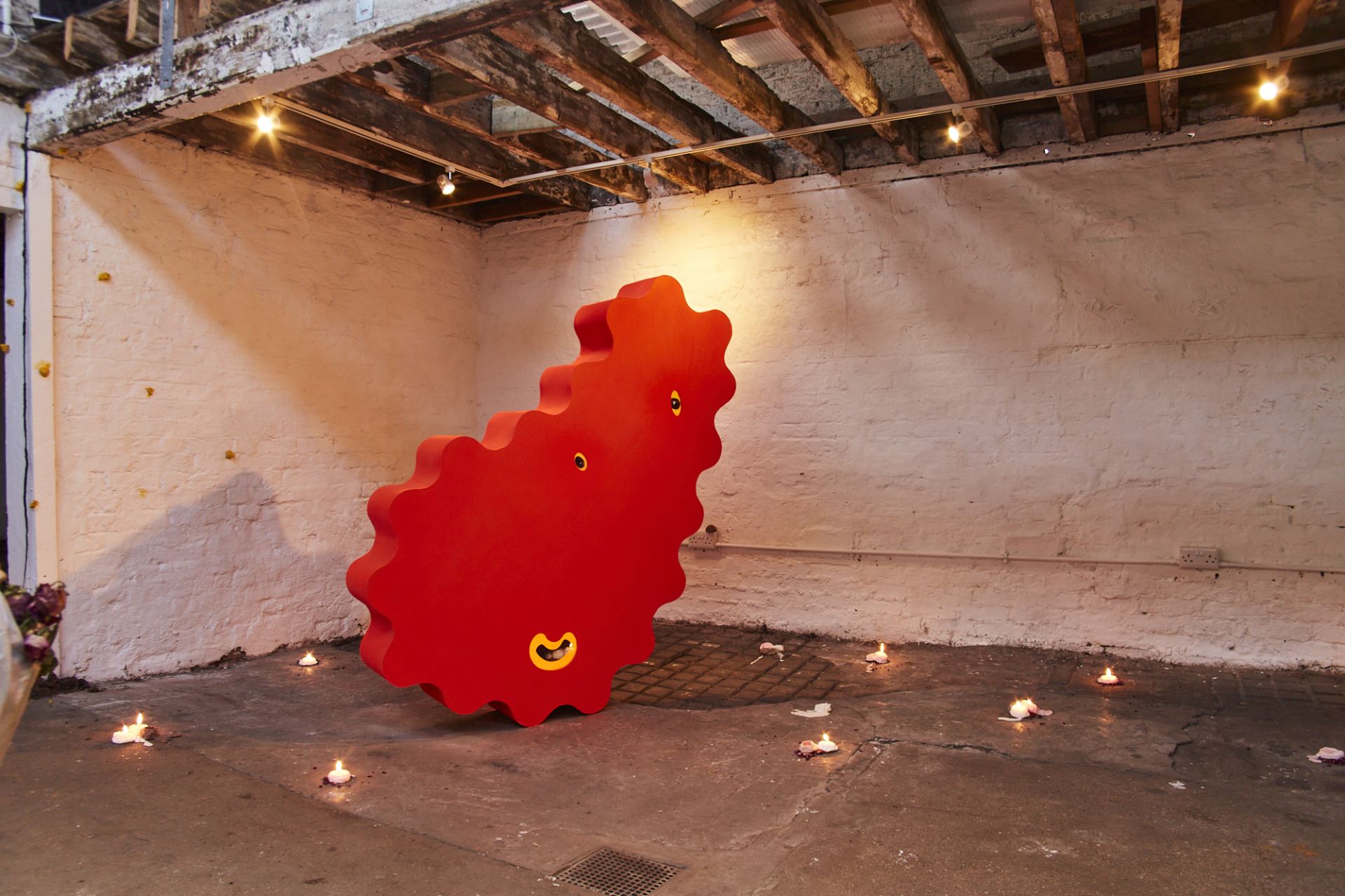 An image of an abstract red sculpture surrounded by candles on the floor of the room around it.