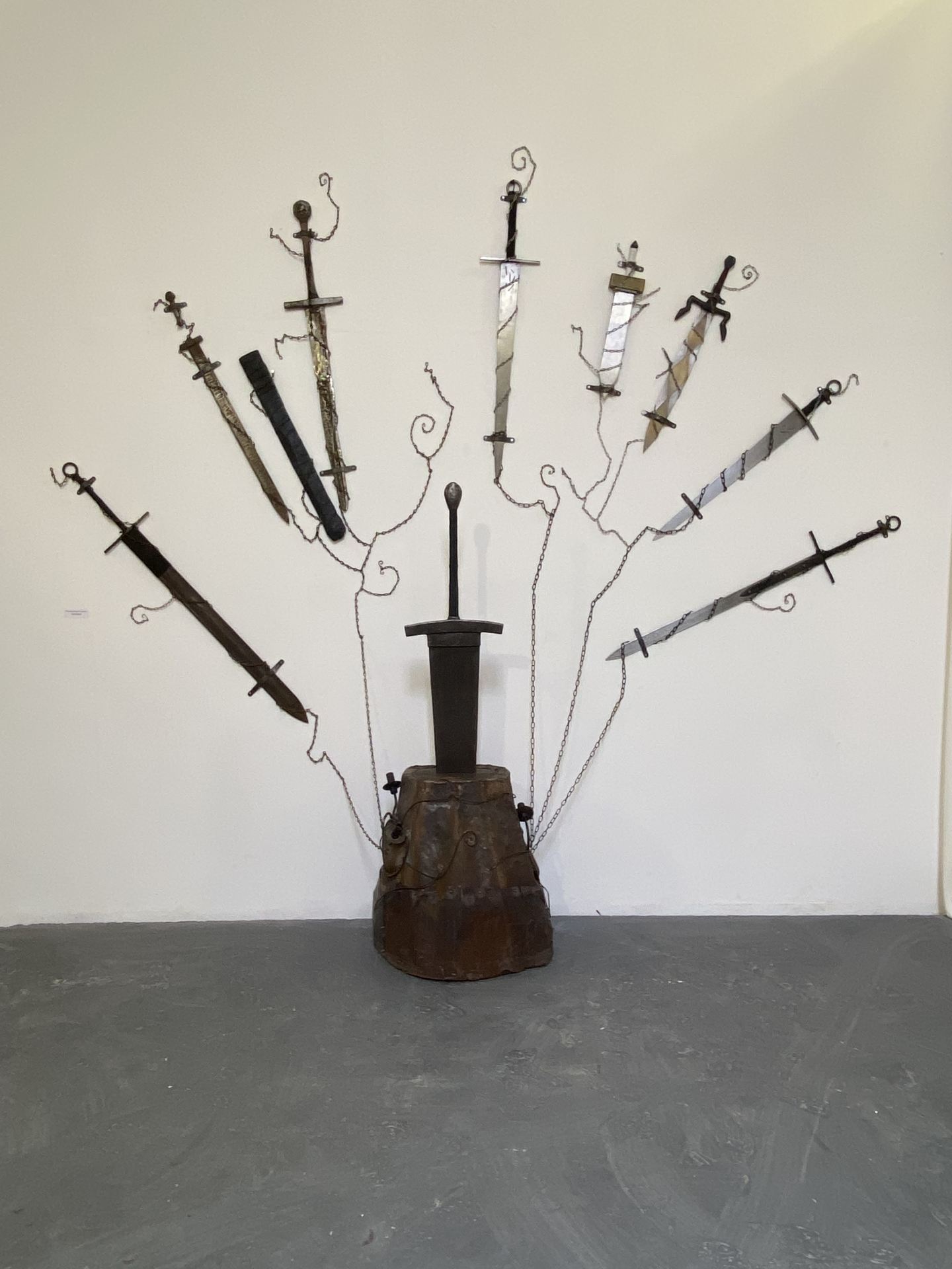 Several swords are wrapped in chains and hang from a white wall. The largest sword emerges  from a brown tree stump in the centre of the composition.
