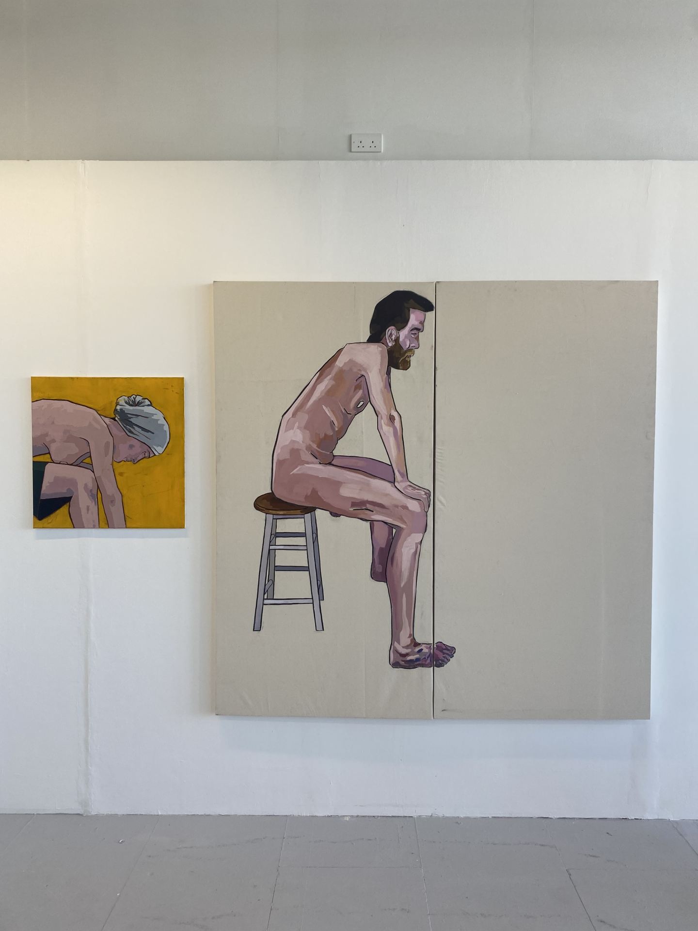 A painting of a person gazing downwards on a yellow canvas, next to a larger diptych of a man sitting naked on a stool.