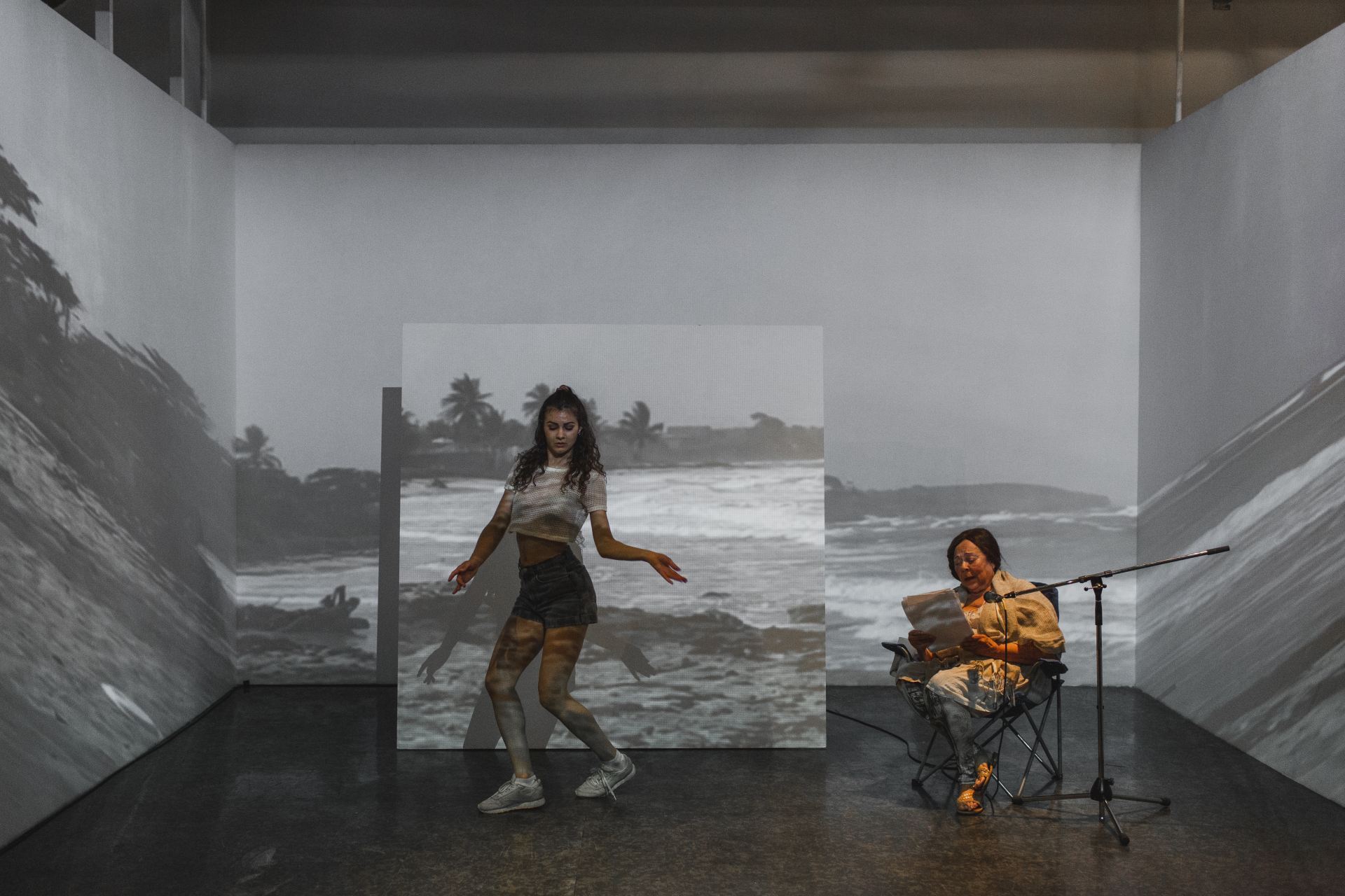 An image of a person dancing and a person sitting and reading. The whole room is covered in a projection of a video of a black and white beach.