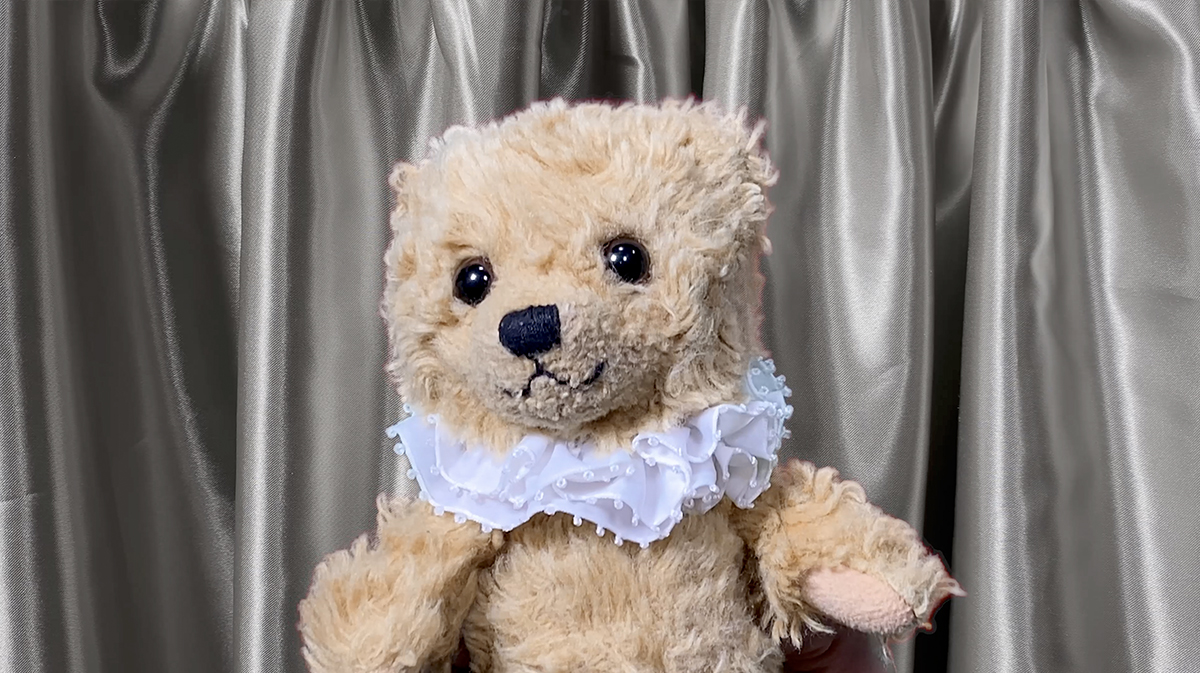 A photo of a teddy bear in front of a silver silk curtain.