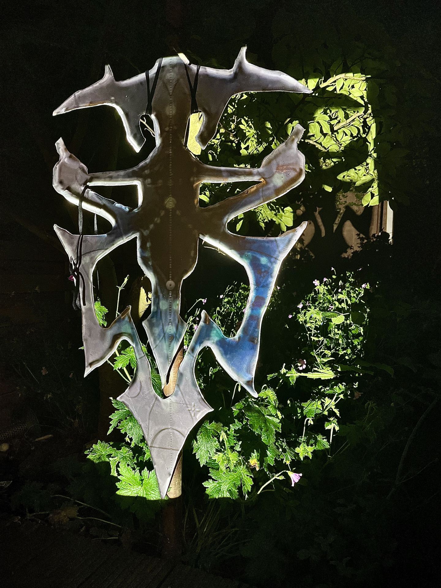 A digitally edited image of a metal sculpture hanging against some plants, set against a black background.
