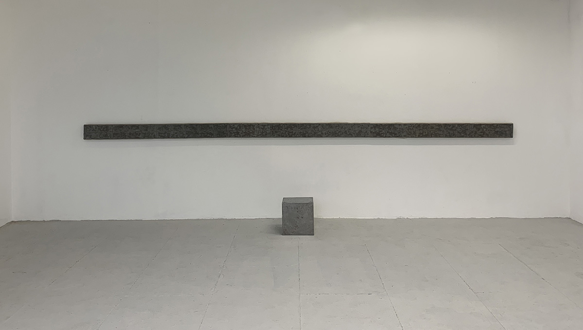 A landscape image of a grey cube beneath a thin, grey painting stretching the width of the wall behind the cube.