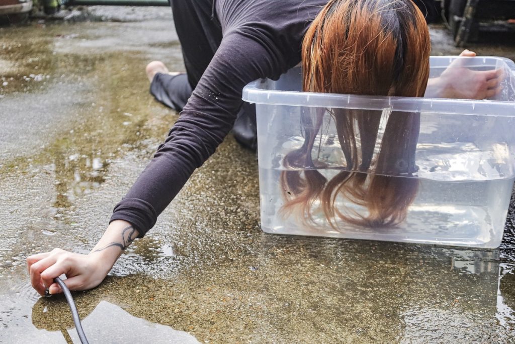 An image of a girl, outside next to a chainlink fence, with her head partially submerged in a transparent plastic box full of water.