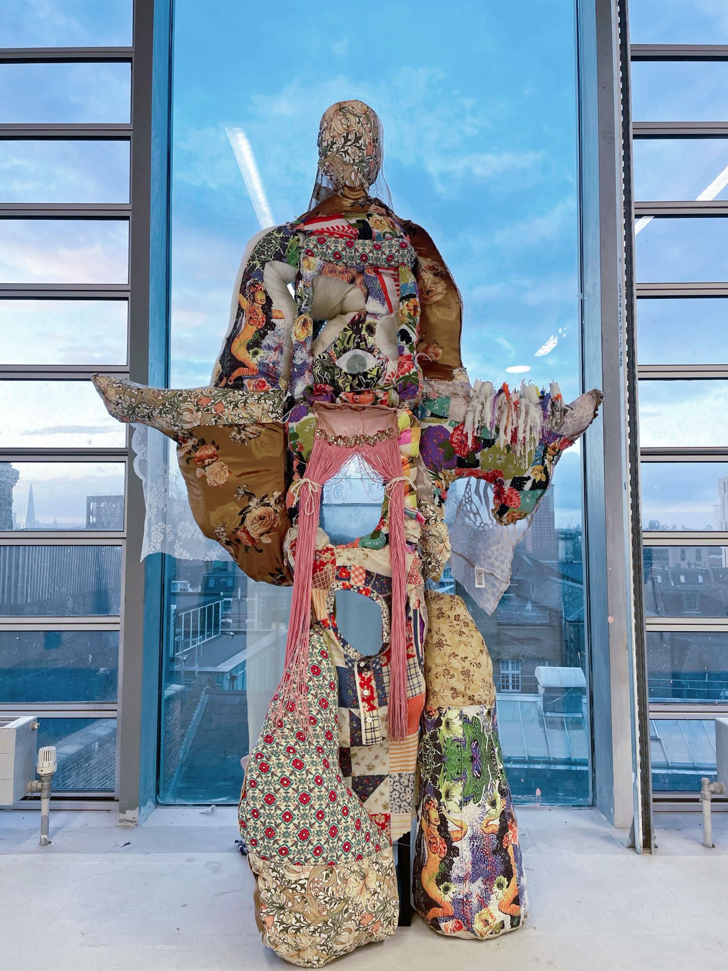 A fabric sculpture in the form of a god with a number of devices for wishing, such as wishing candles, wishing wells and shooting star launchers, against a cityscape seen through a window.