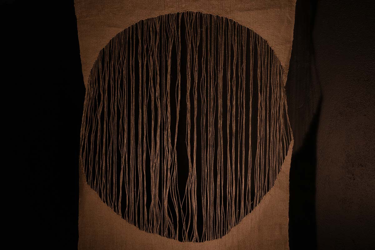 An image of a black circle of negative space, held within a piece of fabric trailing its threads from one end to another.