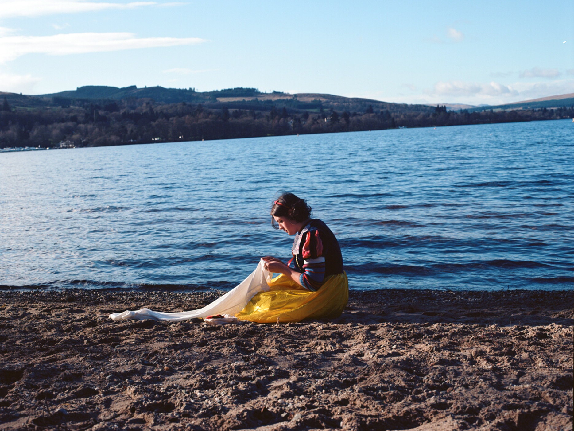 A photo of the Artist dressed as Snow White, sitting and sewing by the edge of Loch Lomond, Scotland.