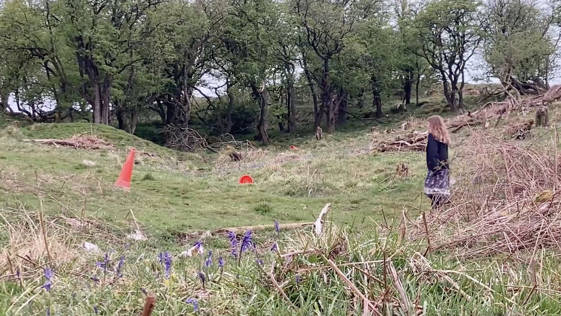 A video still of a person in a field looking at orange traffic cones.