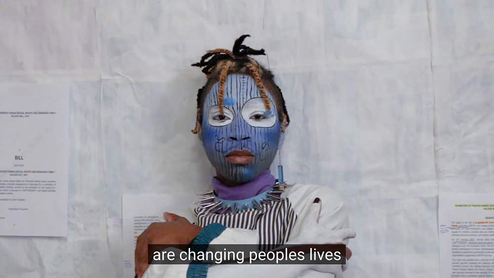 A video still with captioned text. A black person wears blue and white face paint and a blue choker in front of a white background. There are black stripes on their face paint and jacket.
