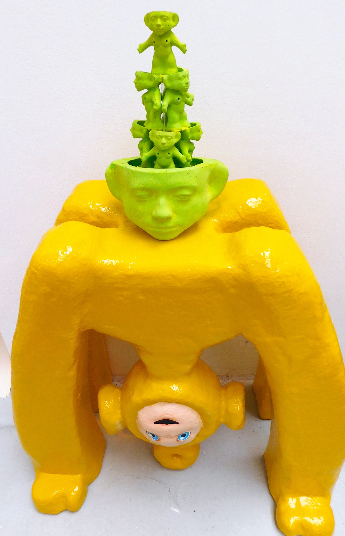 An image of a green stone fountain constructed of trolls sitting on top of a yellow plinth resembling a Teletubby doing a bridge pose. 