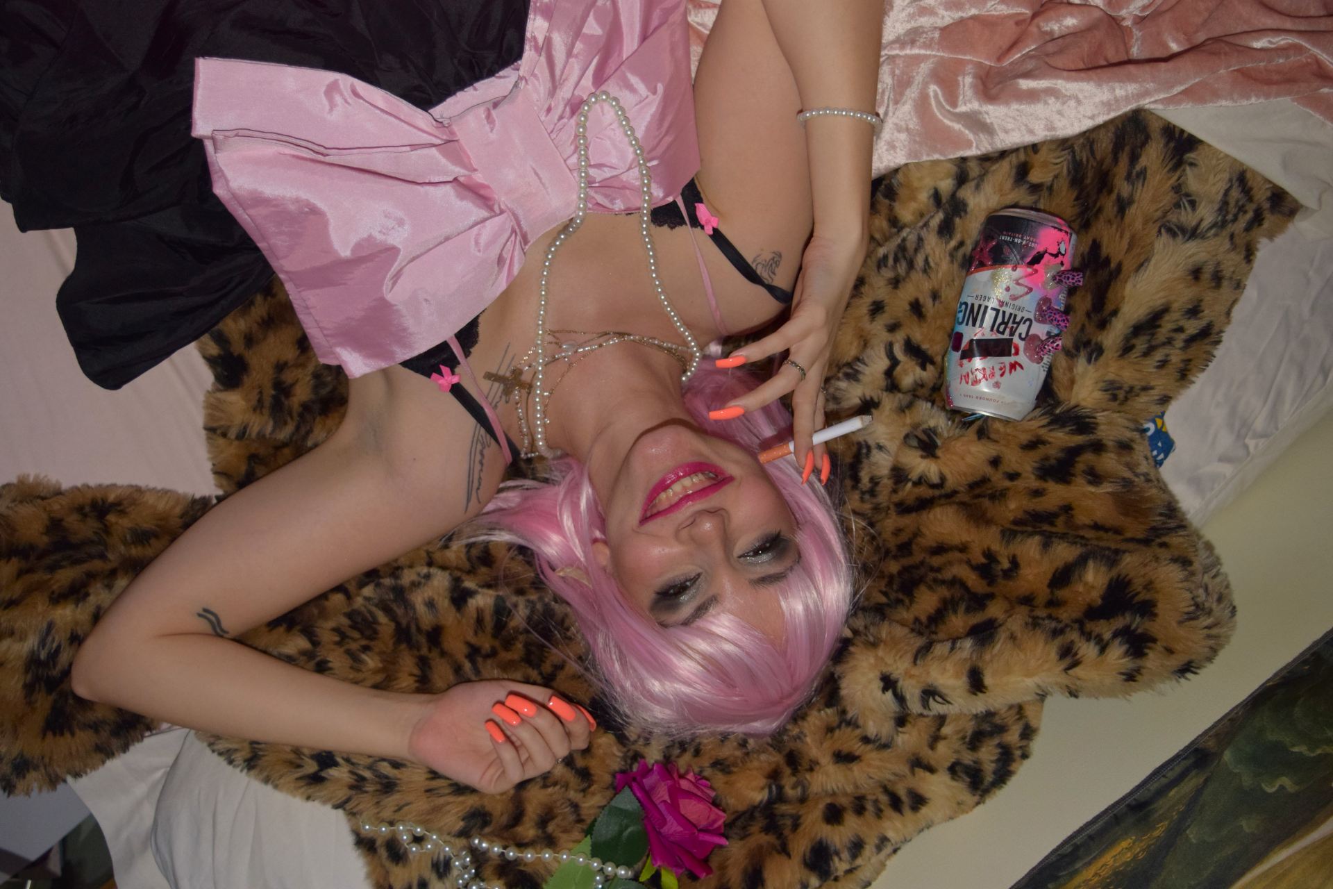 An image of a woman with pink hair, nails and dress laying on a leopard skin rug, smoking.