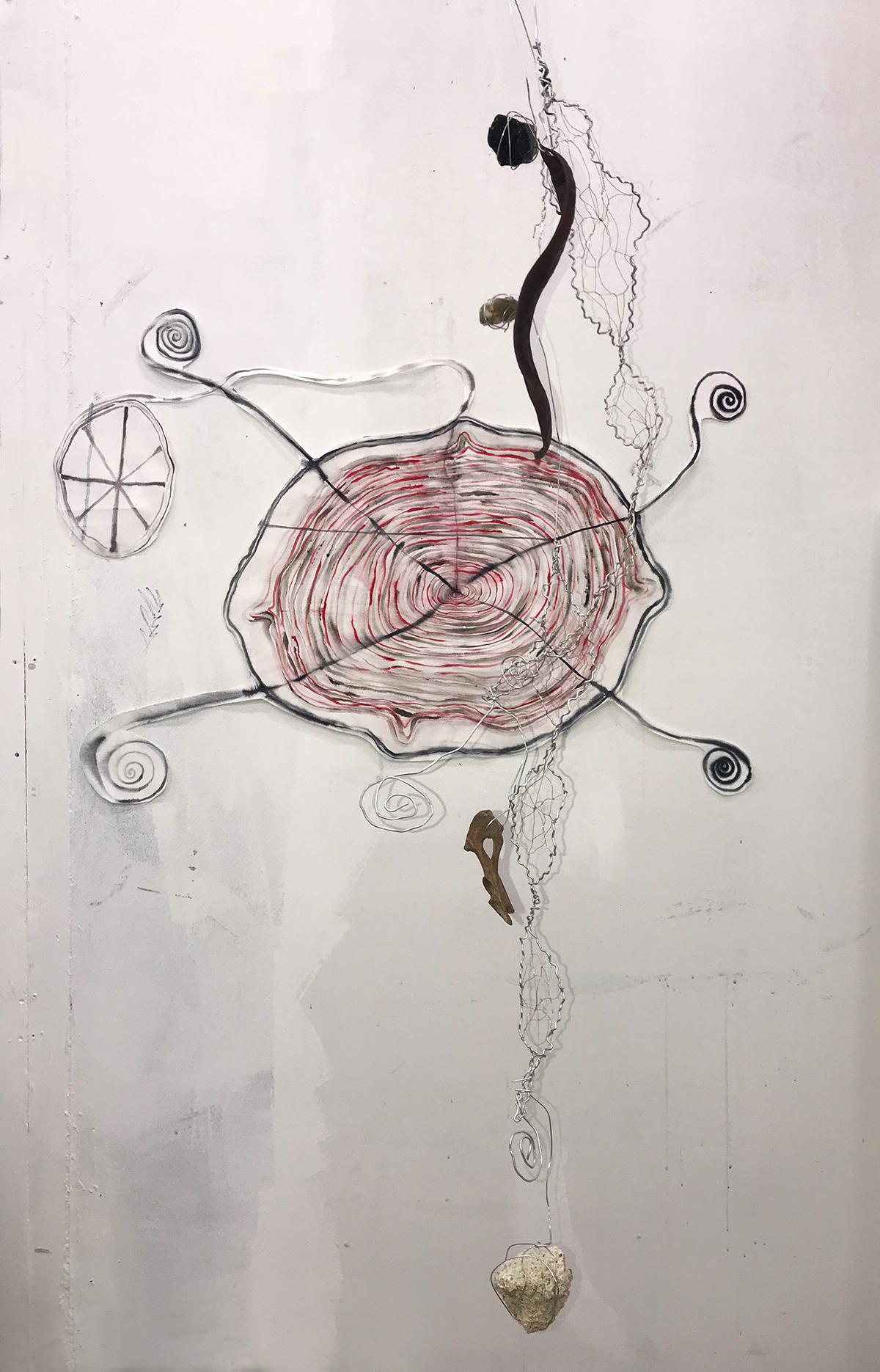 A black and red ink spiral with a cross leading to small outer spirals, in front hangs a weblike wire sculpture which holds pieces of coral, bone, tooth and obsidian.