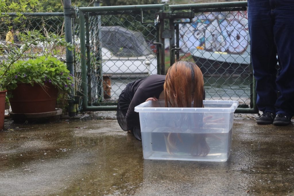 An image of a girl, outside next to a chainlink fence, with her head partially submerged in a transparent plastic box full of water.
